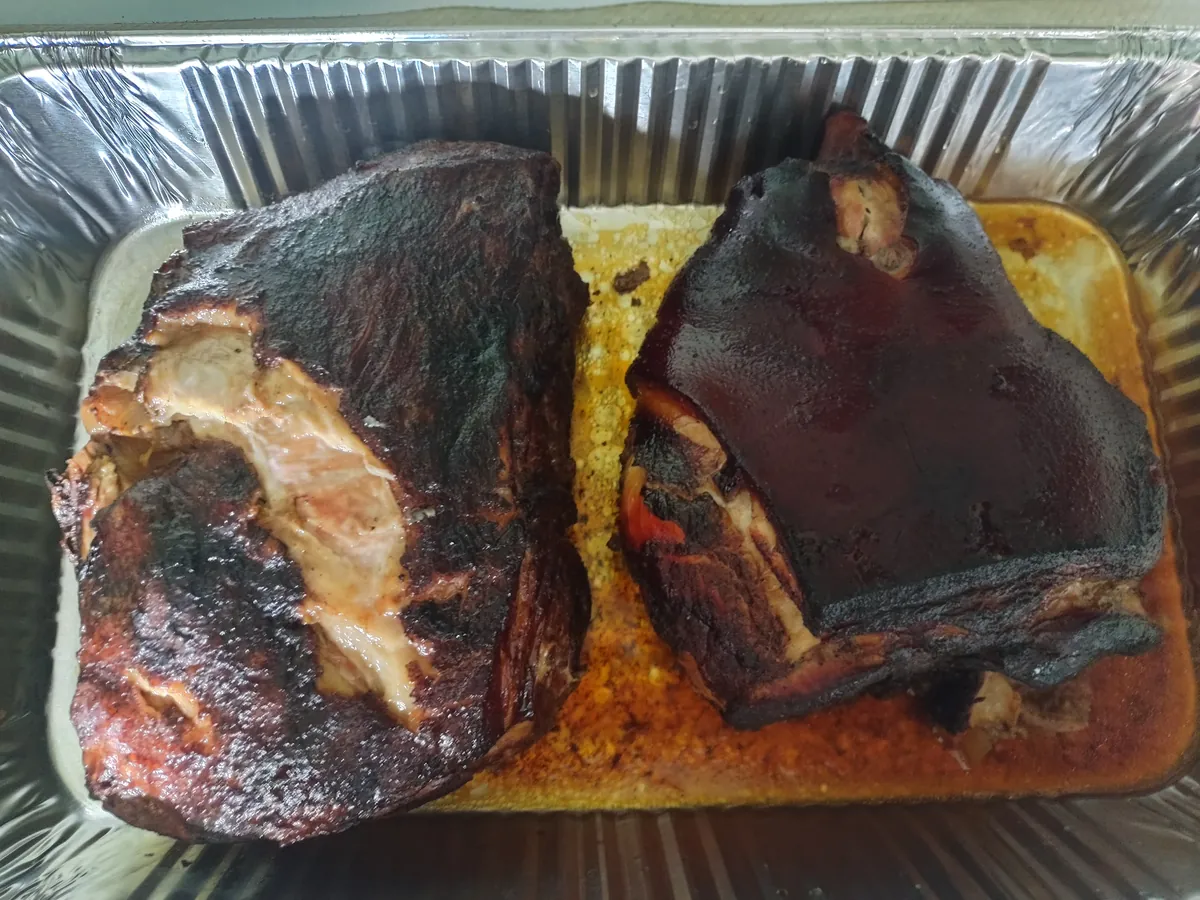 Two whole smoked pork butts with dark bark in a foil pan